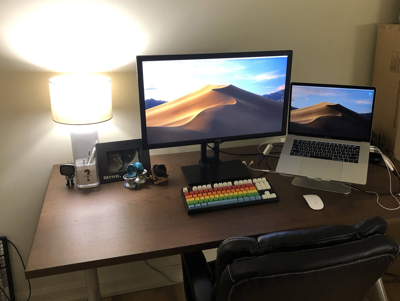A large wooden desk with a laptop, a monitor, a rainbow keyboard, and some desk toys